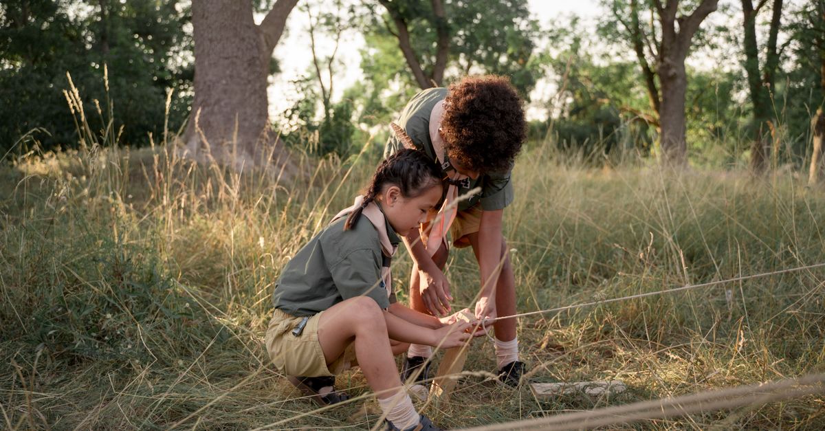 Two young scouts tying knots in a grassy campsite, focused on teamwork and learning.